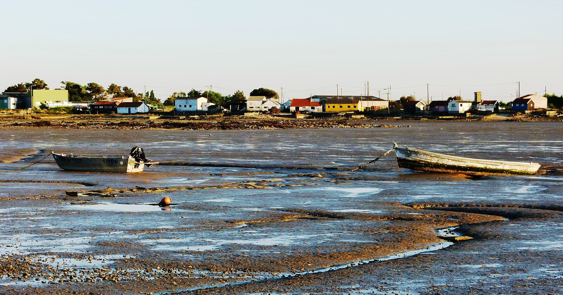 Estran découvert à marée basse au coucher du soleil à Bourcefranc-le-Chapus, deux barques échouées sur la vase et la ligne des maisons basses du village au bord du rivage.
