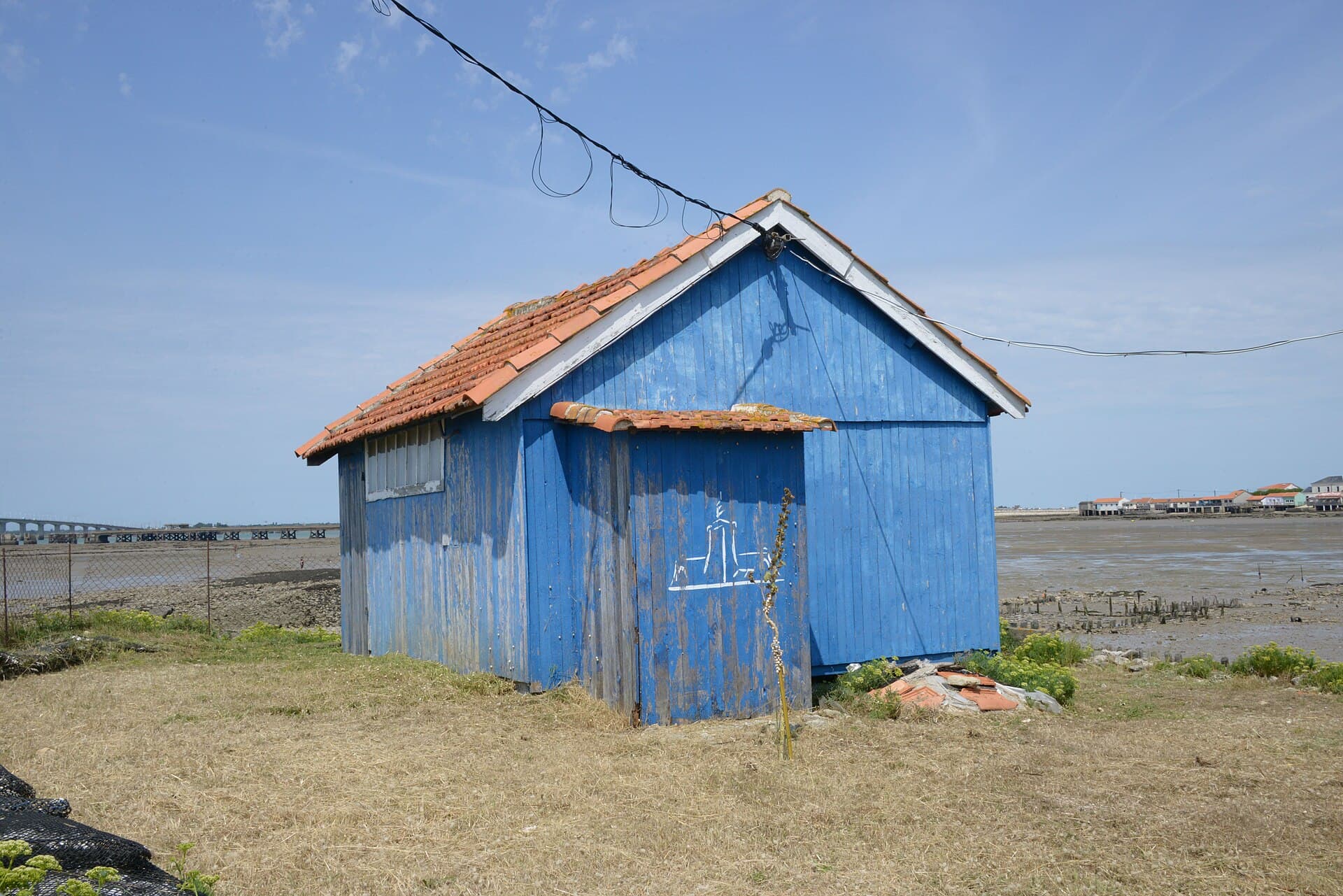 Cabane ostréicole en bois bleu au toit de tuiles, posée au bord des vasières du bassin de Marennes-Oléron, avec le viaduc d'Oléron à l'horizon