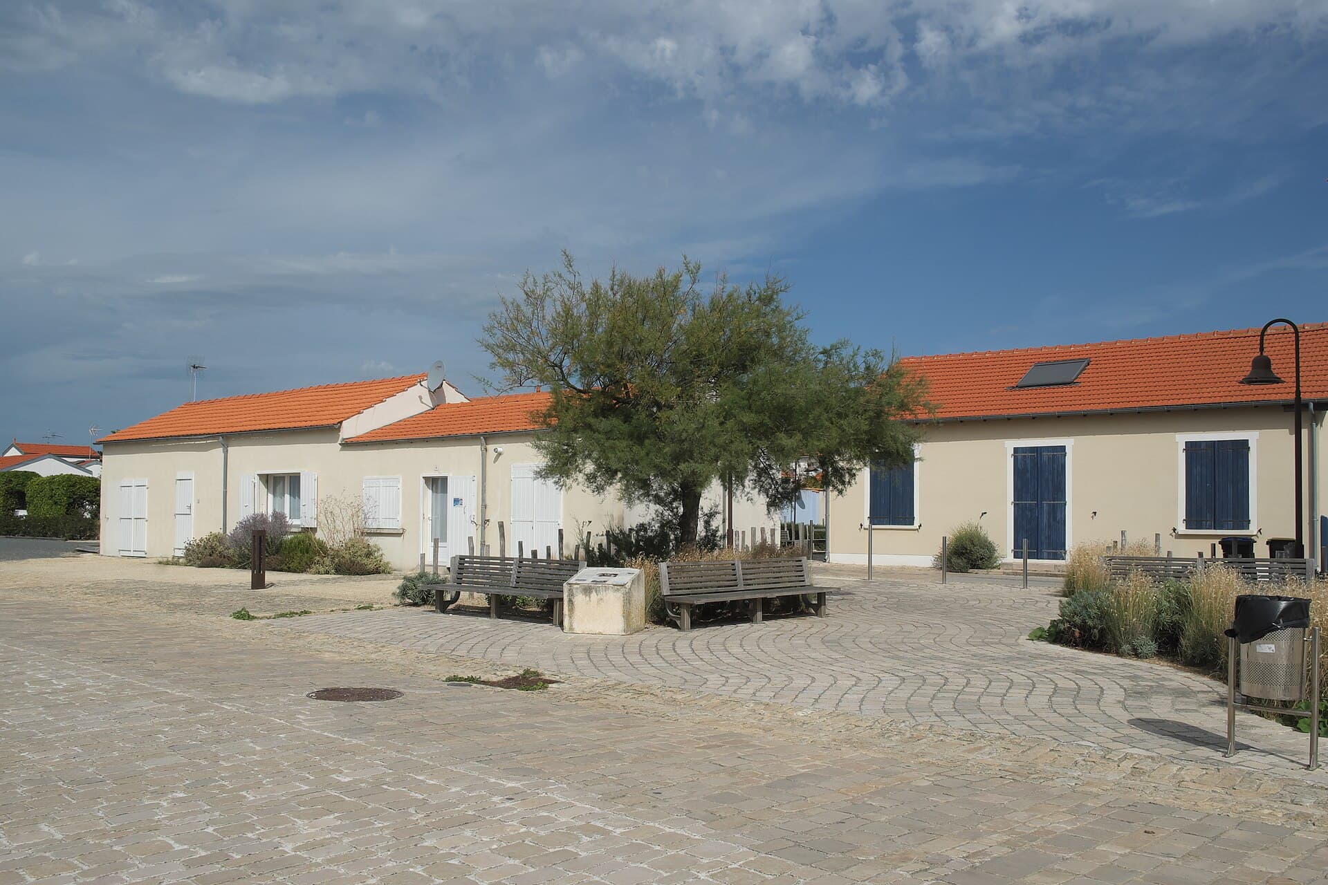 Petites maisons blanches à toiture de tuiles orange et volets bleus, autour d'une place pavée plantée d'un tamaris, à Châtelaillon-Plage.