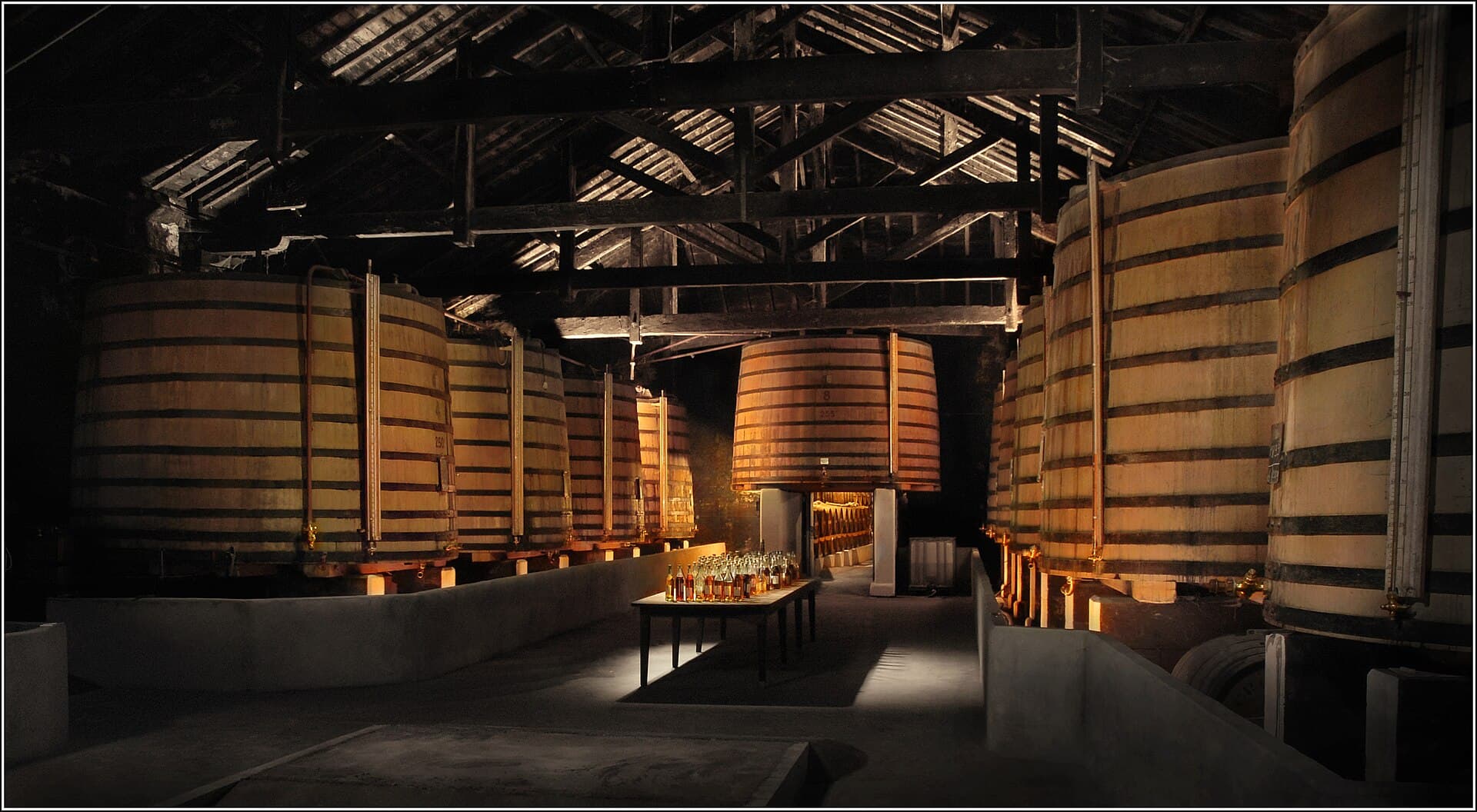 Intérieur d'un chai charentais : de grands foudres de chêne cerclés alignés de chaque côté sous une charpente ancienne, éclairés d'une lumière chaude.