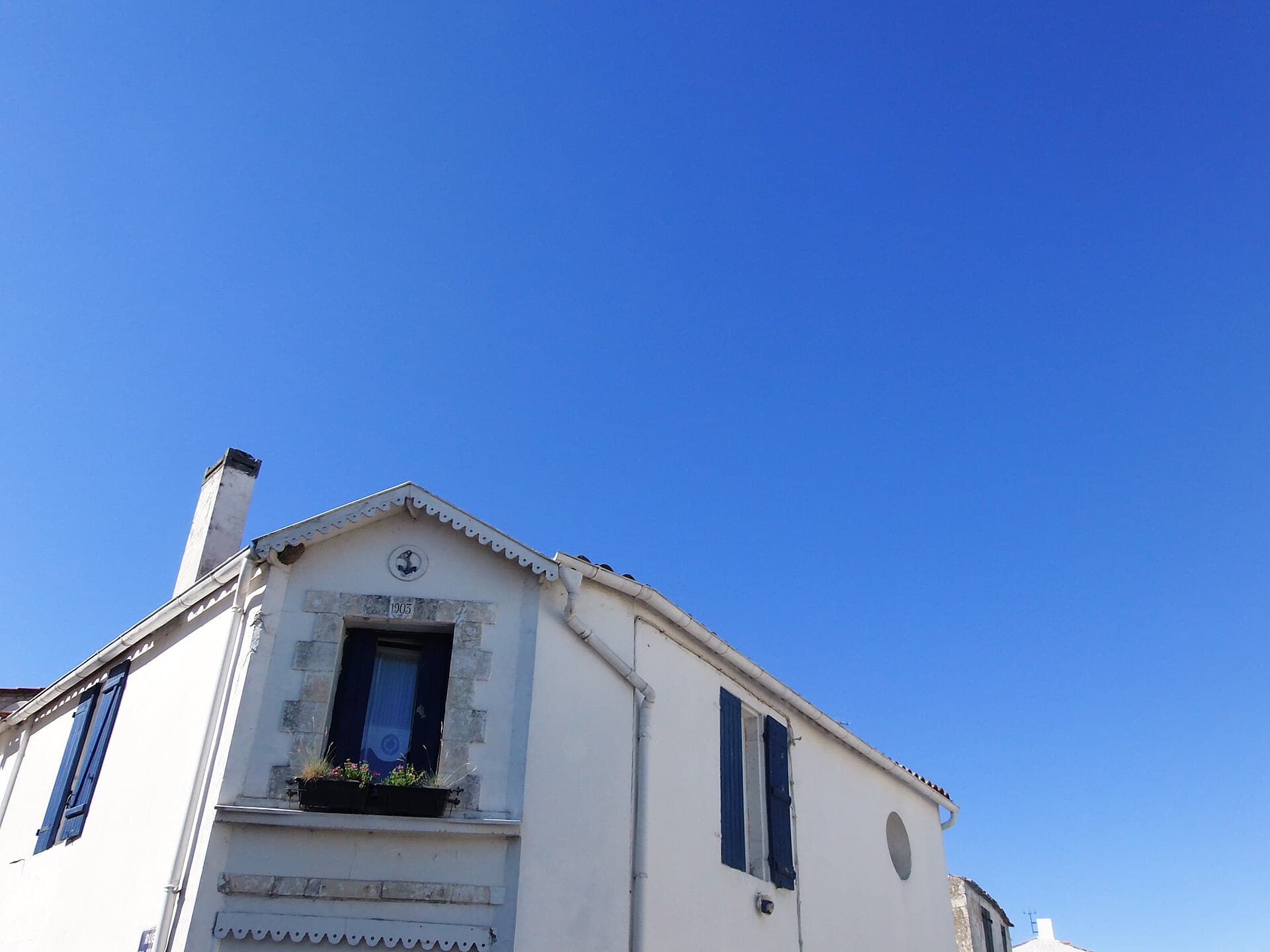 Pignon blanc d'une maison de bourg à Dolus-d'Oléron, volets bleus, encadrement de pierre et jardinière fleurie, sous un ciel bleu profond