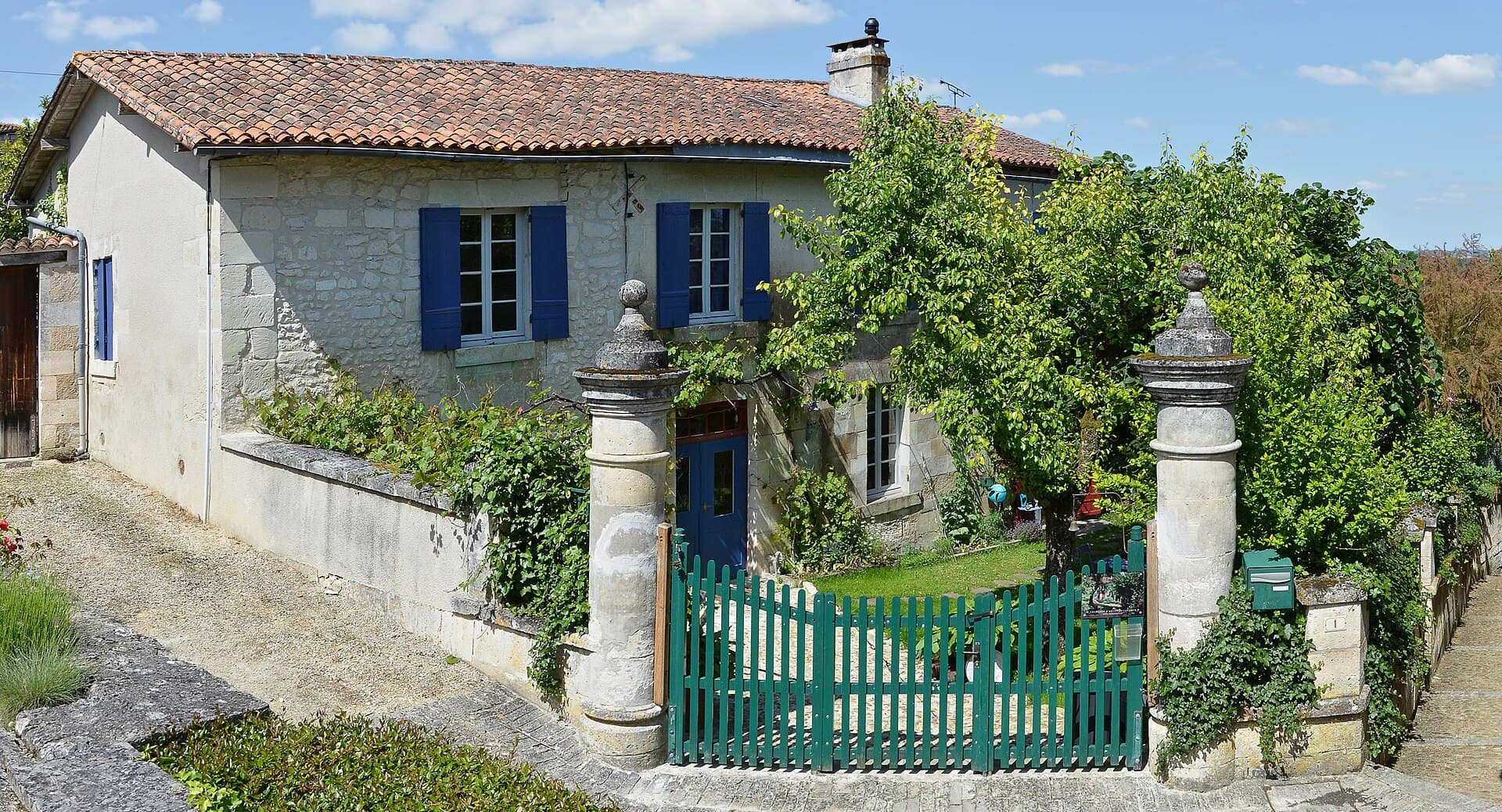 Maison charentaise en pierre au toit de tuiles, volets bleus et portail vert, entourée de verdure sous un ciel d'été