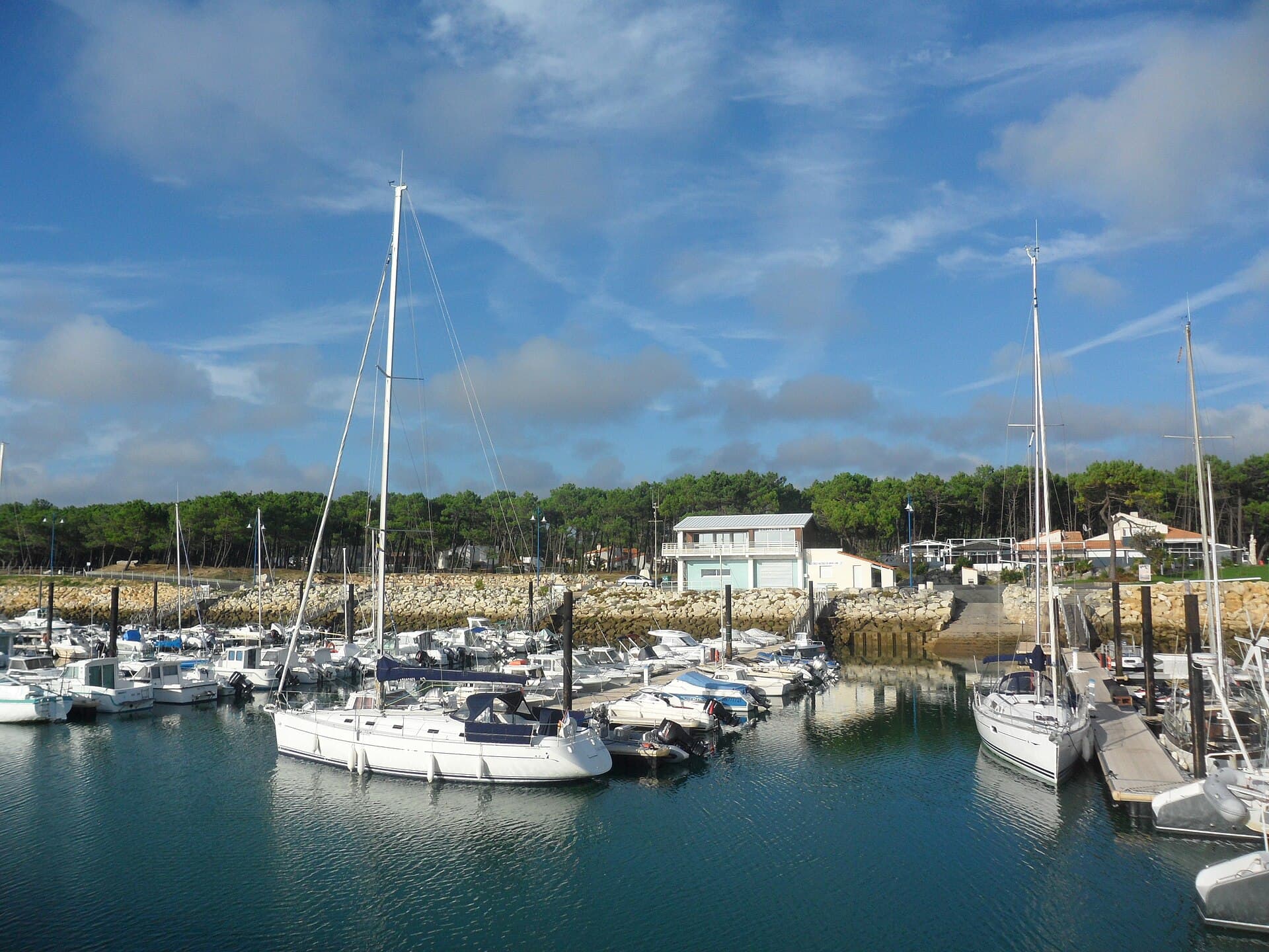 Le port de plaisance de La Palmyre, ses voiliers amarrés devant la pinède et les maisons du bord de mer, sous un ciel bleu