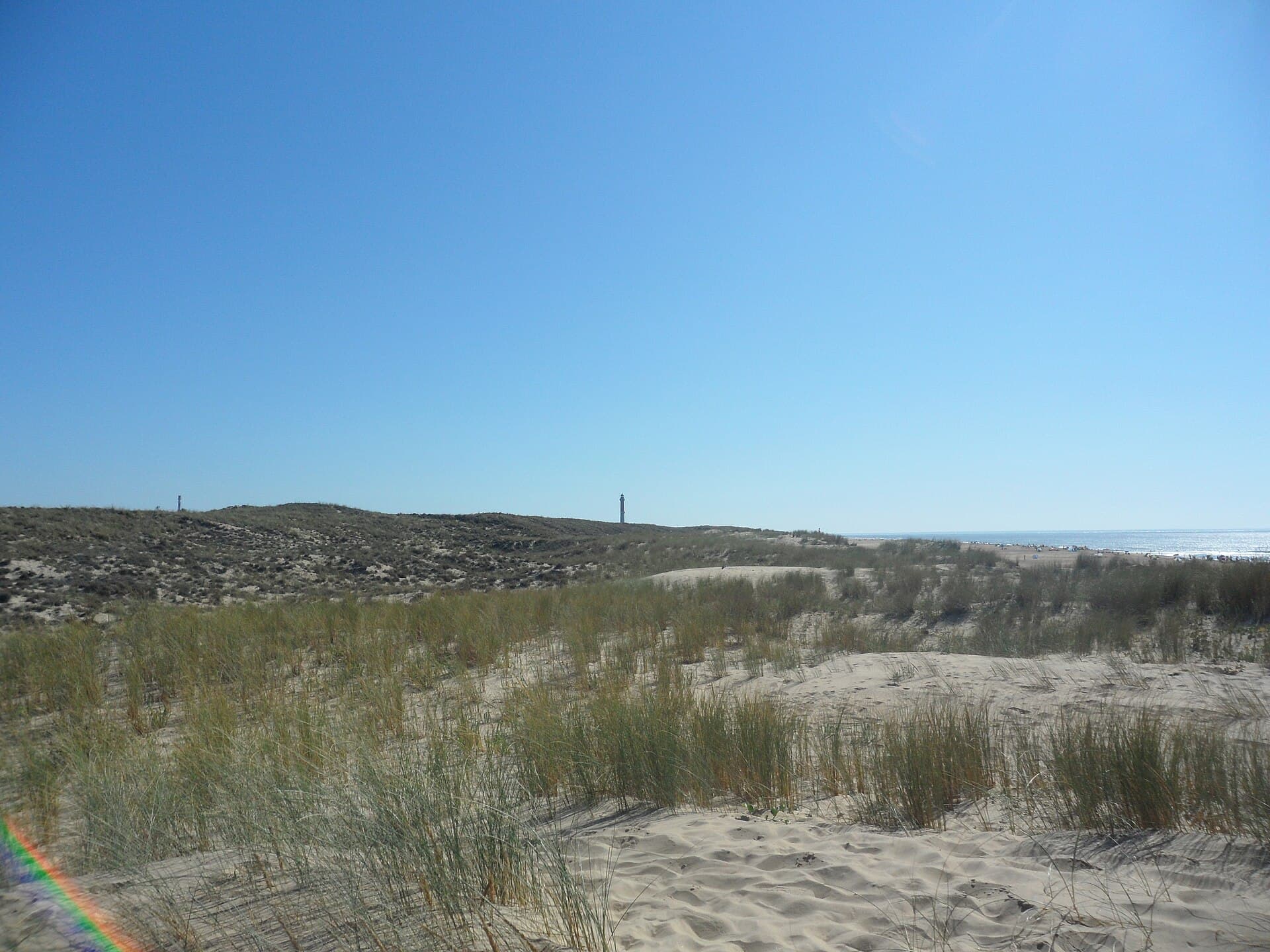 Dunes couvertes d'oyats descendant vers l'océan sous un ciel bleu, avec la silhouette d'un phare au loin