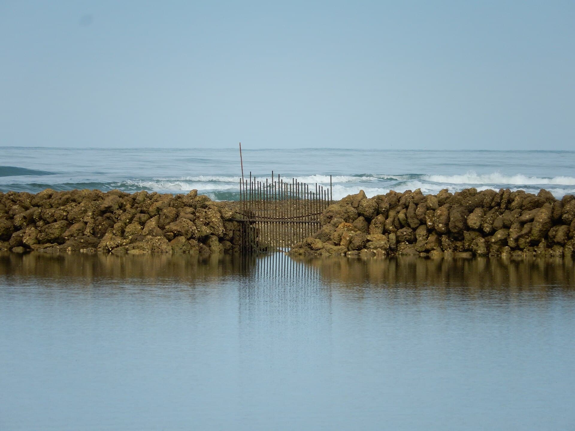 Écluse à poissons en pierre sur l'estran de l'île d'Oléron, ses claies de bois au centre et les vagues de l'océan qui déferlent à l'horizon.