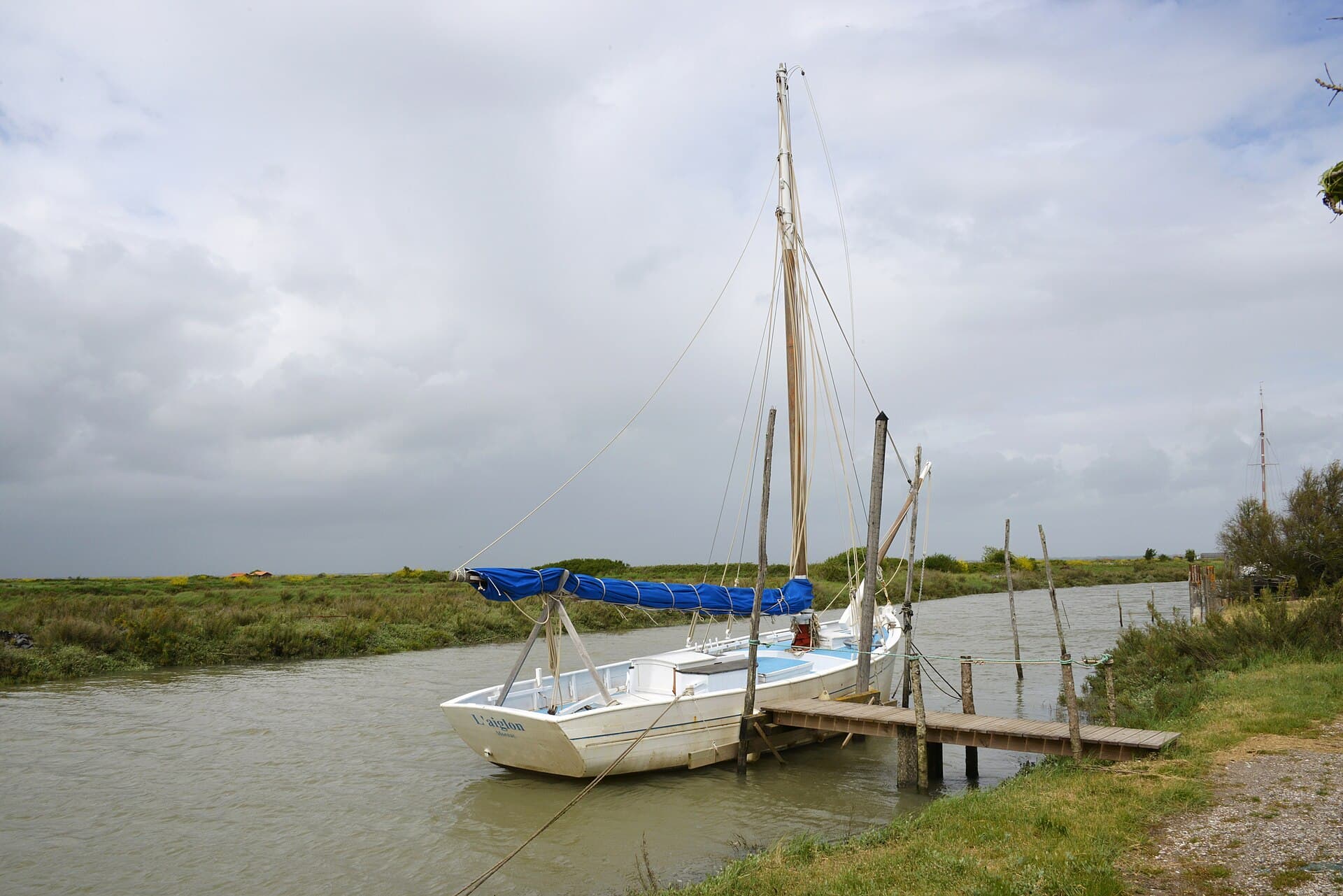 Un vieux sloop ostréicole blanc amarré à un ponton de bois sur un chenal bordé de marais, sous un ciel nuageux