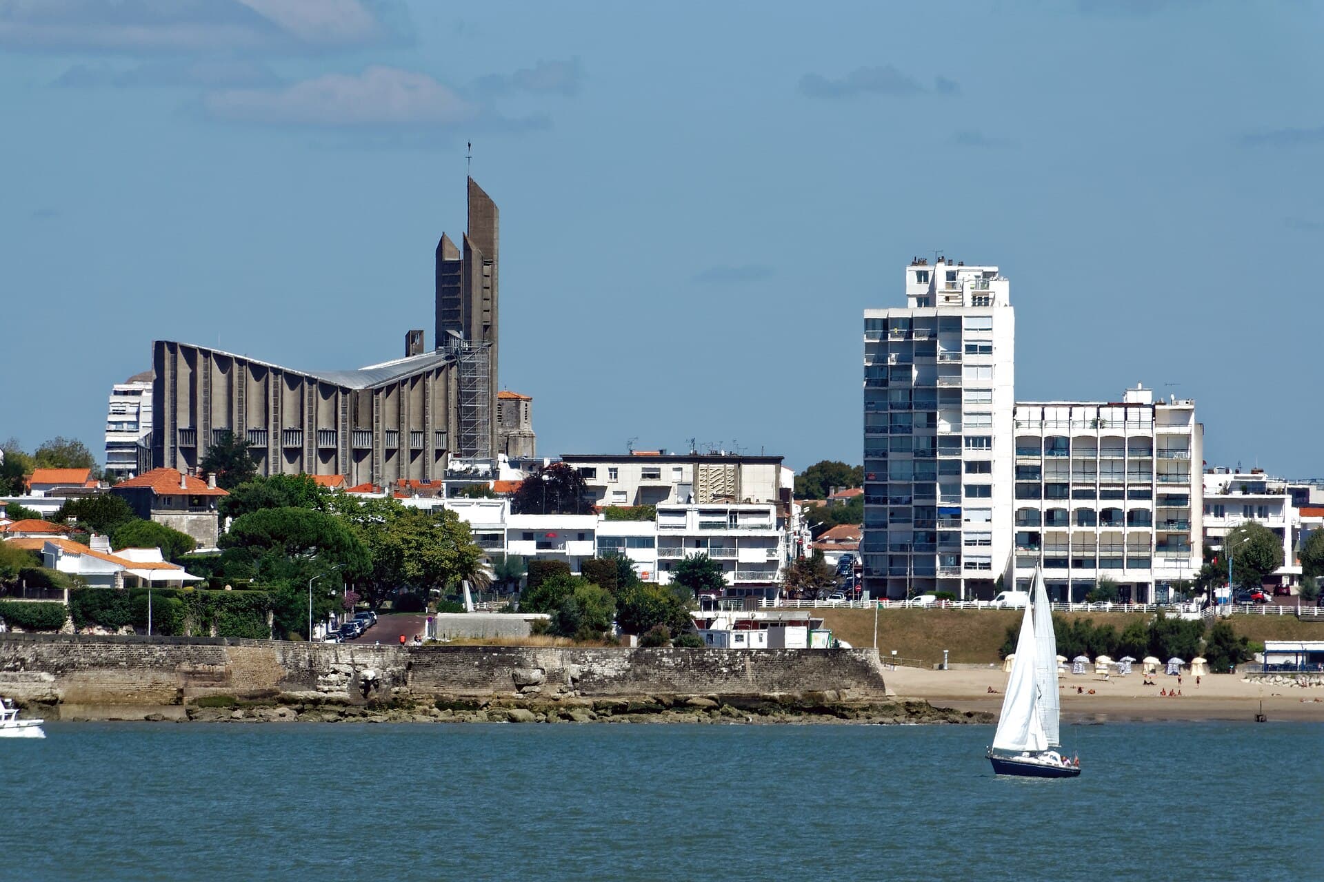 Le front de mer de Royan vu depuis l'eau, avec ses villas à toits de tuiles, ses immeubles blancs, la plage et un voilier au premier plan