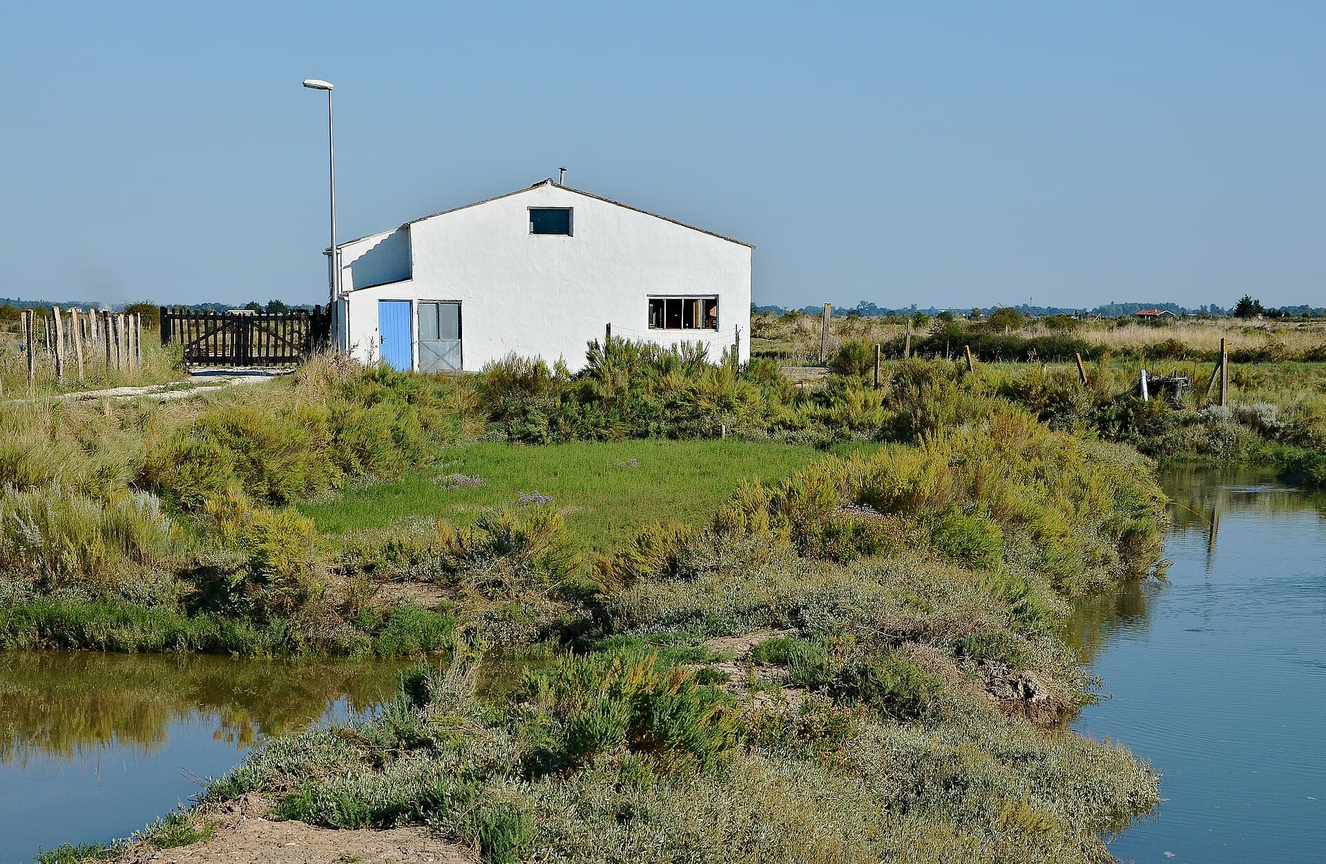 Maison blanche à porte bleue au bord d'un chenal du marais de la Seudre, entourée de végétation basse sous un ciel bleu